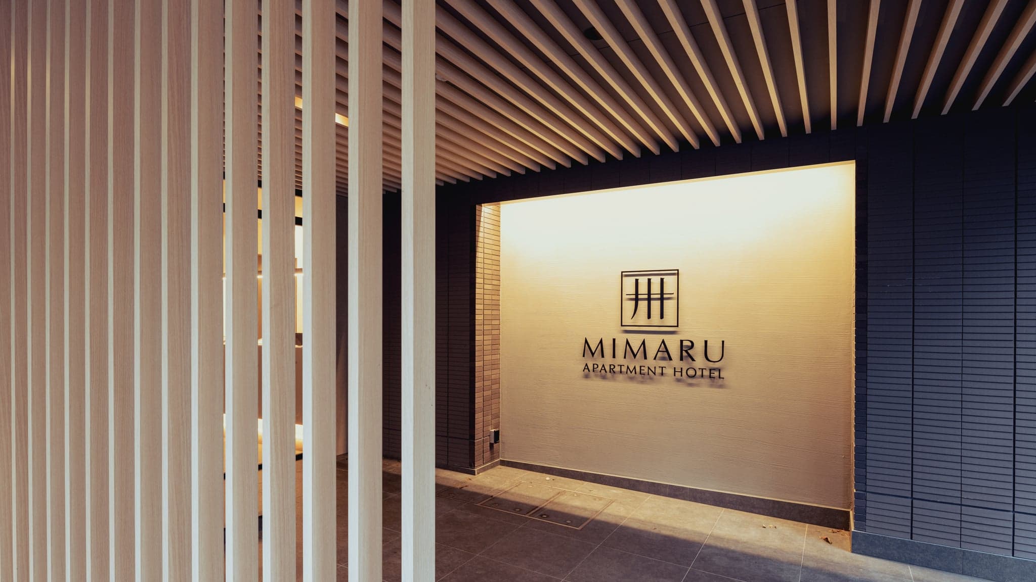 The entrance of a MIMARU apartment hotel with its logo on a lit wall