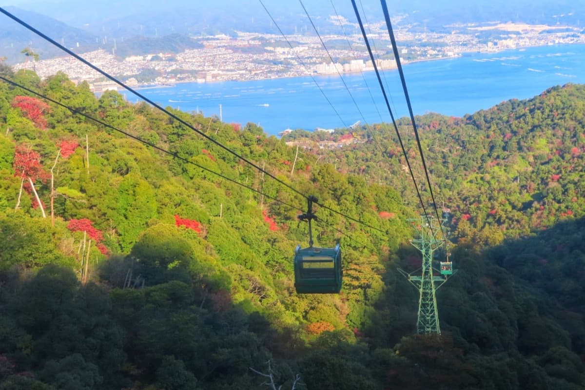 A small Miyajima Ropeway gondola above a forested slope, with the Inland Sea and Hiroshima's coast in the distance