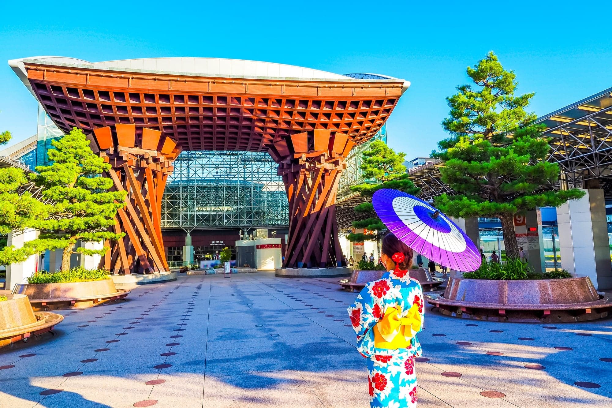 The wooden Tsuzumi Gate in front of Kanazawa Station on a clear day, with a visitor in a yukata holding a purple parasol