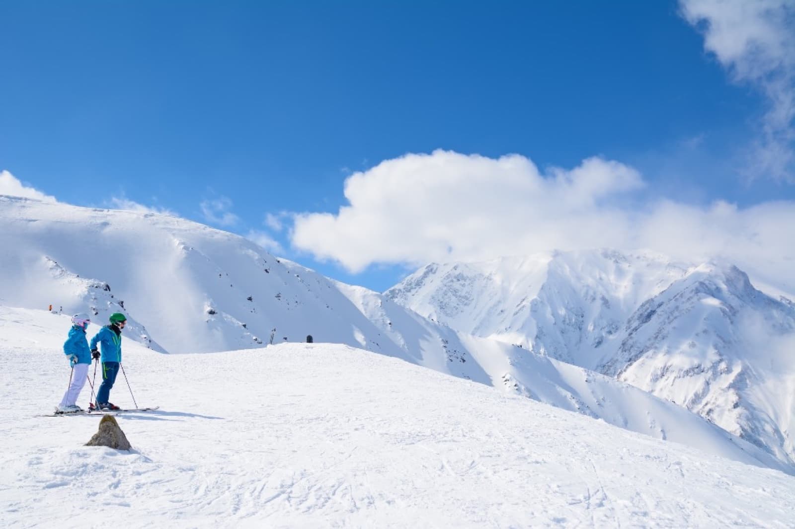 Two skiers on a snowy ridge looking out over snow-covered mountain peaks under a clear blue winter sky