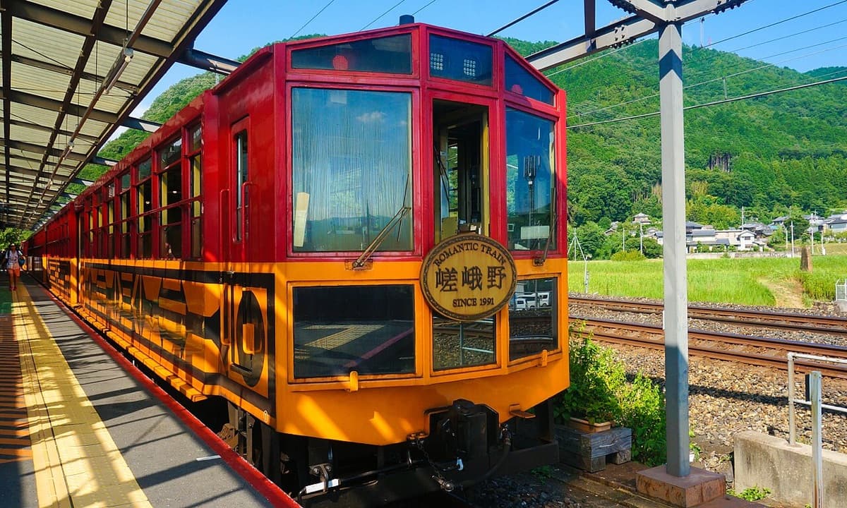 The red and orange carriages of the Sagano Romantic Train standing at a covered platform with green hills behind