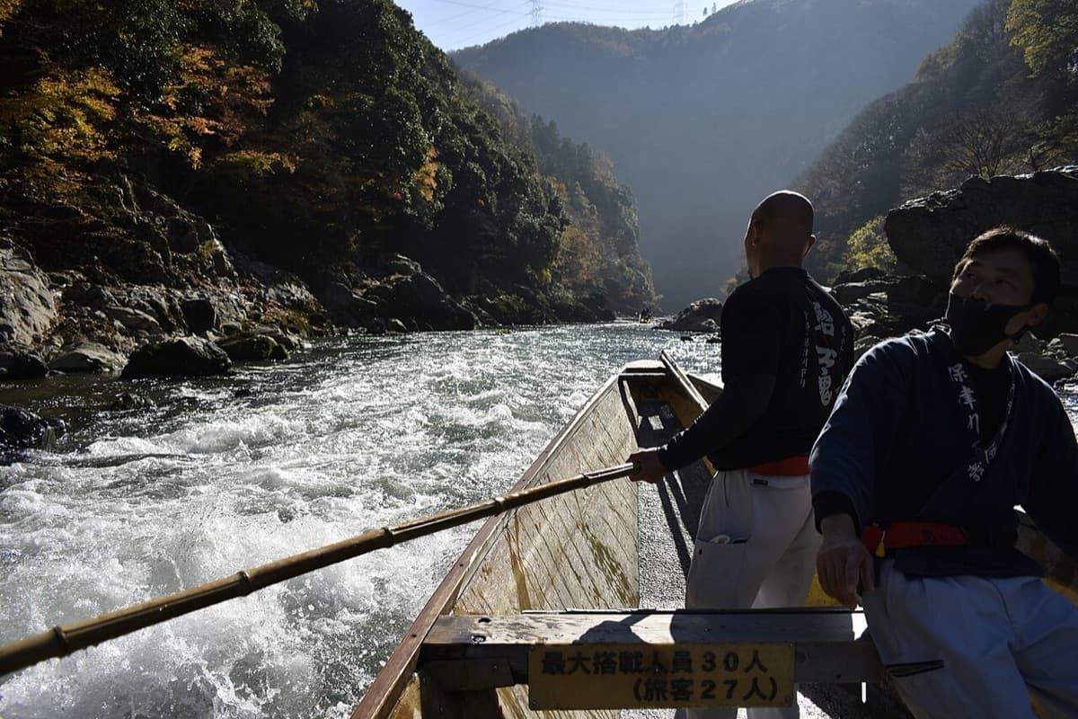 Boatmen steering a wooden Hozugawa river boat with long poles through white water in a wooded gorge