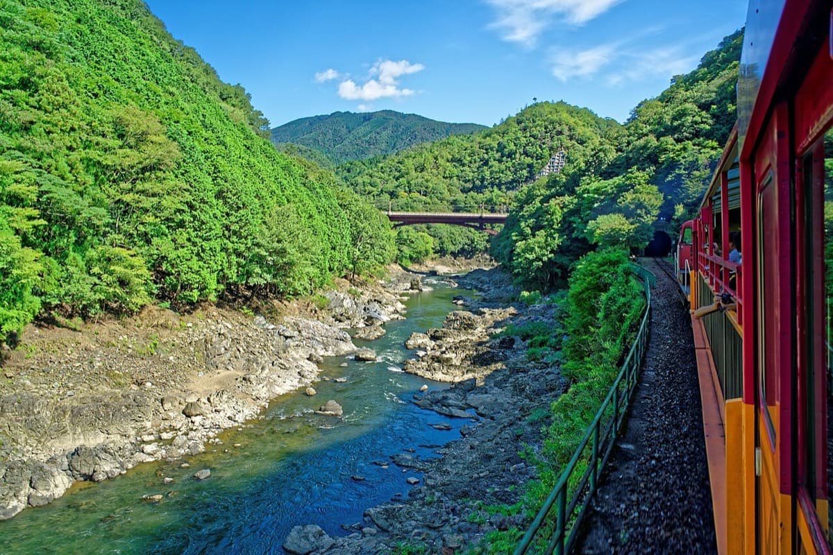 View from the Sagano train of the Hozu River gorge: a rocky river between steep green hillsides, with a bridge in the distance