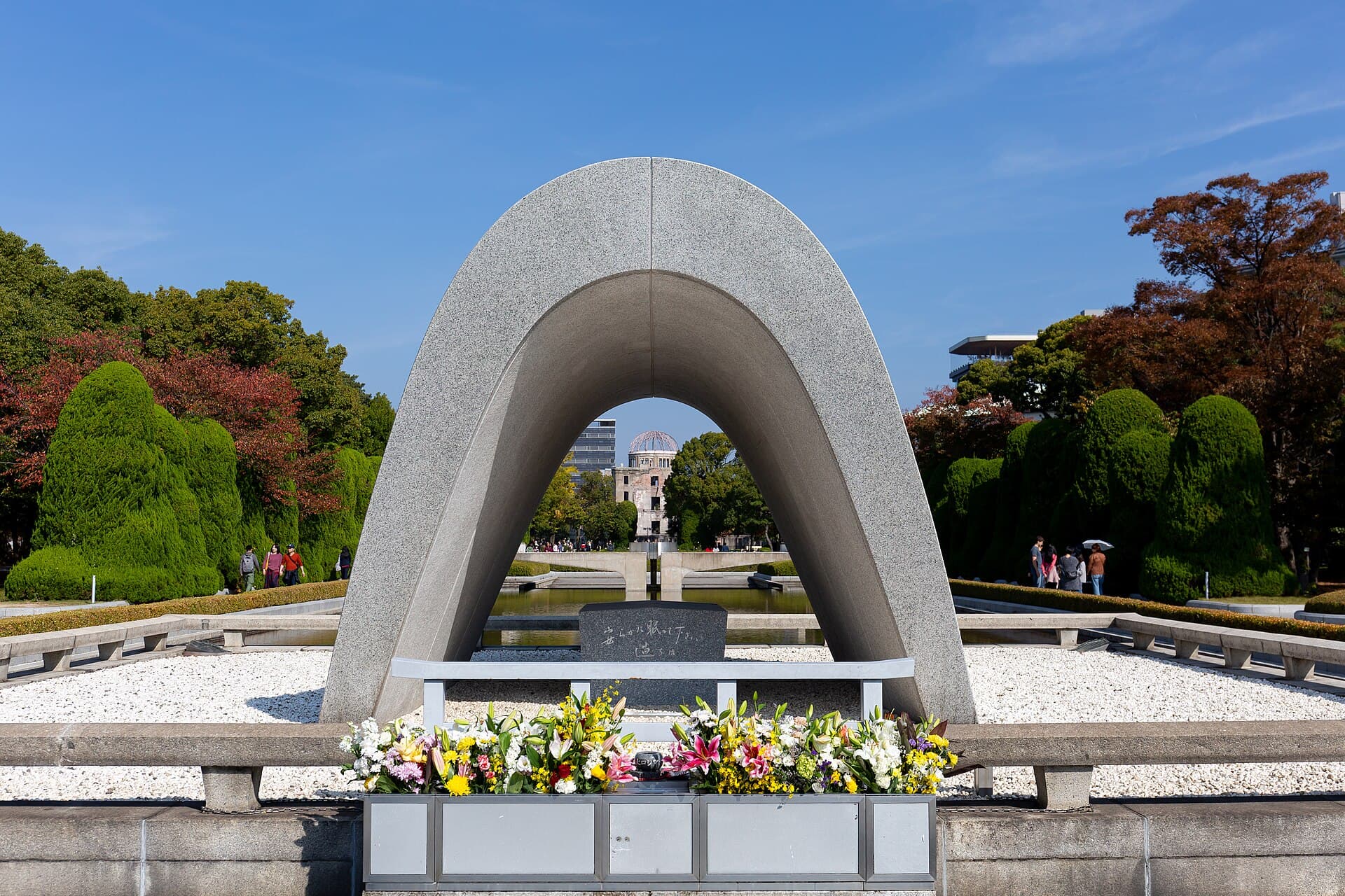 The arched stone Cenotaph in Peace Memorial Park with flowers in front, framing the Atomic Bomb Dome in the distance