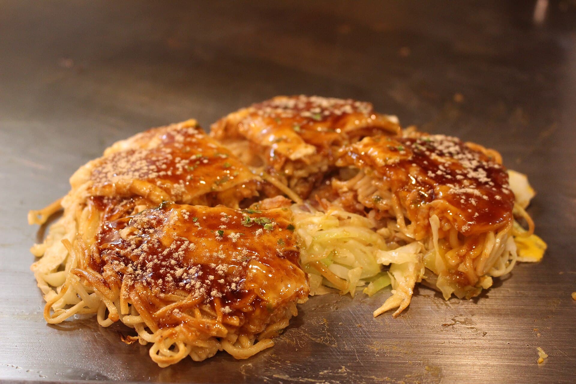 A Hiroshima-style okonomiyaki with a layer of noodles, brushed with dark sauce and sprinkled with seaweed and sesame, on a steel griddle