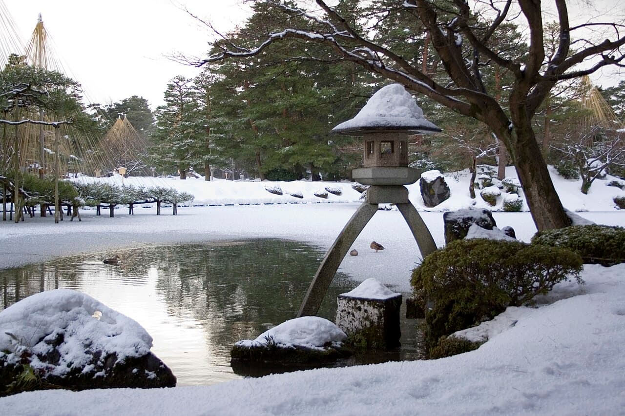 Kenrokuen's two-legged stone lantern beside a pond, with snow on the lantern, rocks and trees