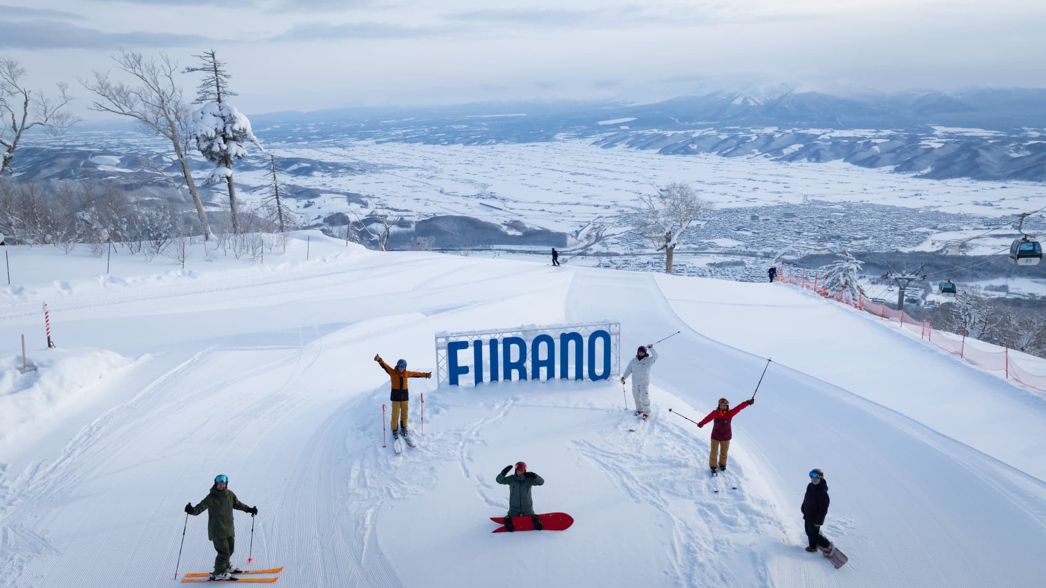 A group of skiers and a snowboarder posing by the big FURANO sign on a slope above the snowy Furano basin