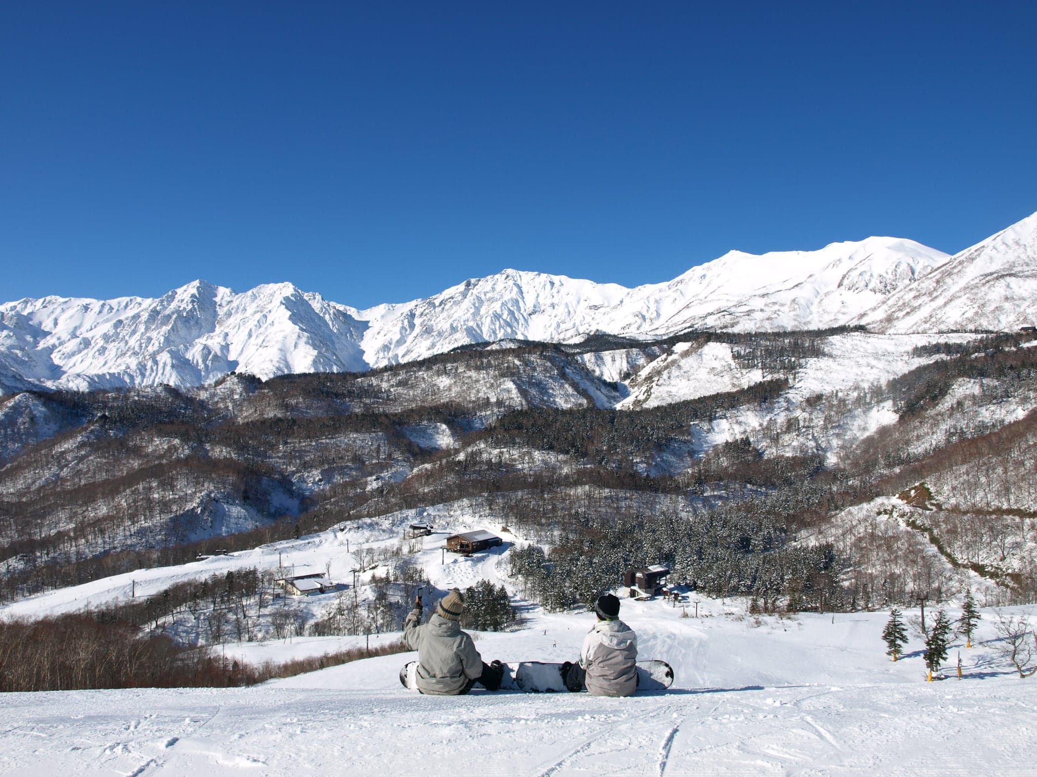 Two snowboarders sitting on a sunny slope at Tsugaike in Hakuba, with the snow-covered Northern Alps behind them