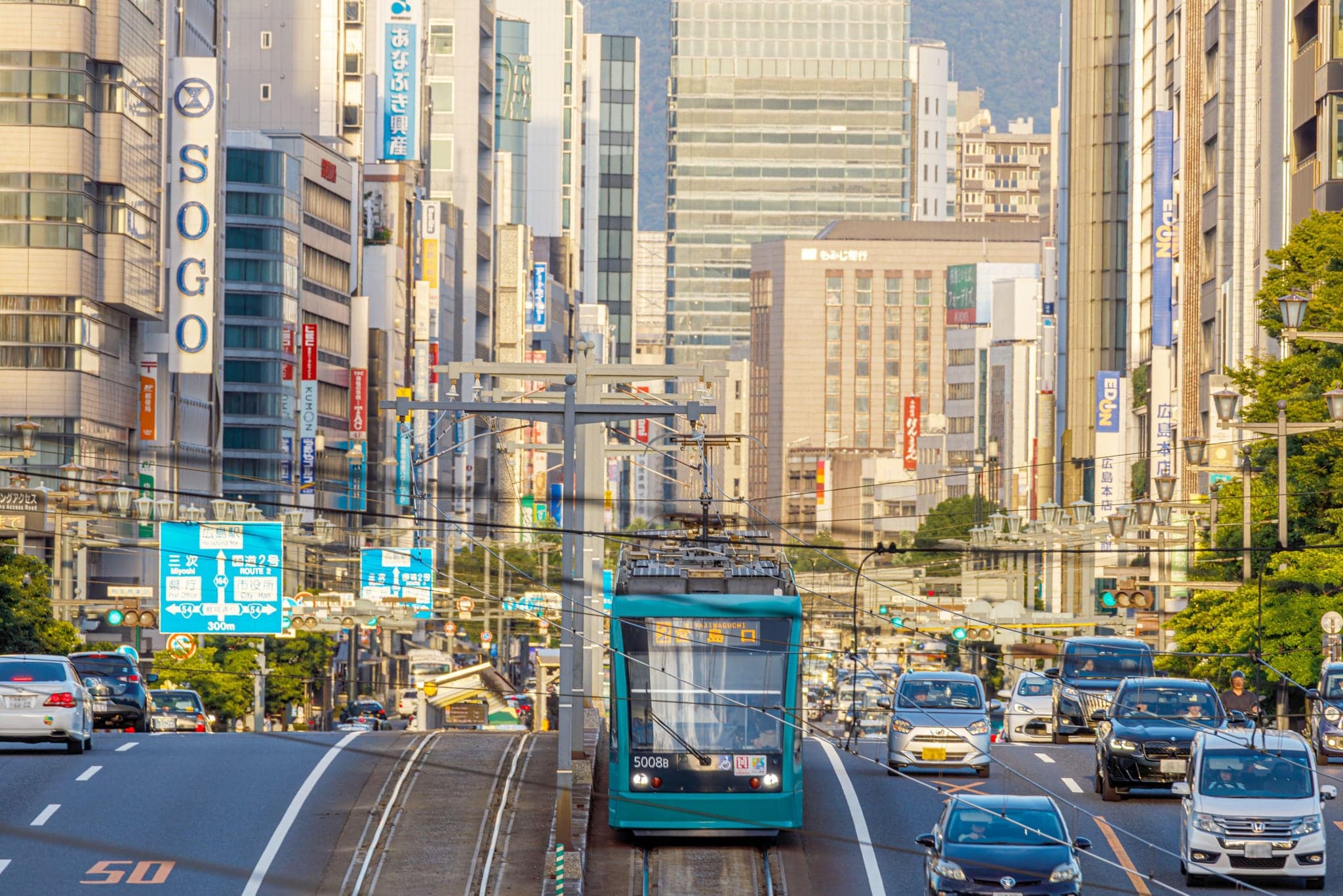 A green-and-white Hiroden streetcar on a wide avenue in central Hiroshima, with cars and city buildings around it