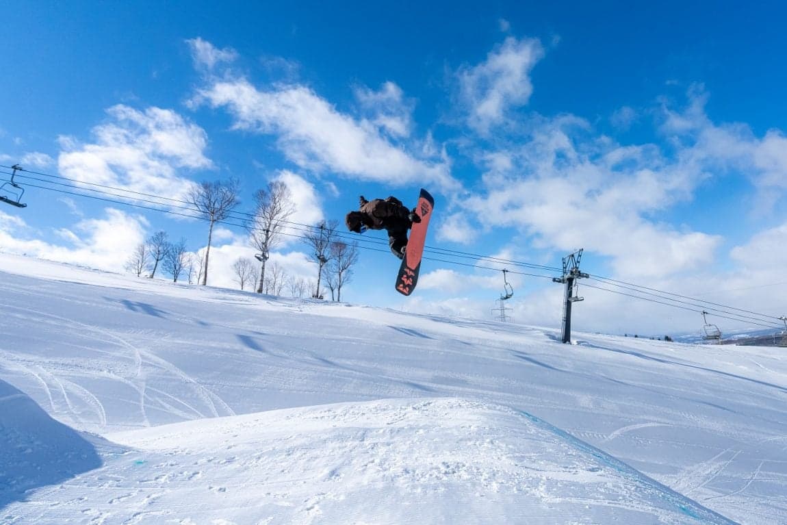 A snowboarder grabbing in the air over a kicker at Rusutsu under a blue sky, a chairlift behind
