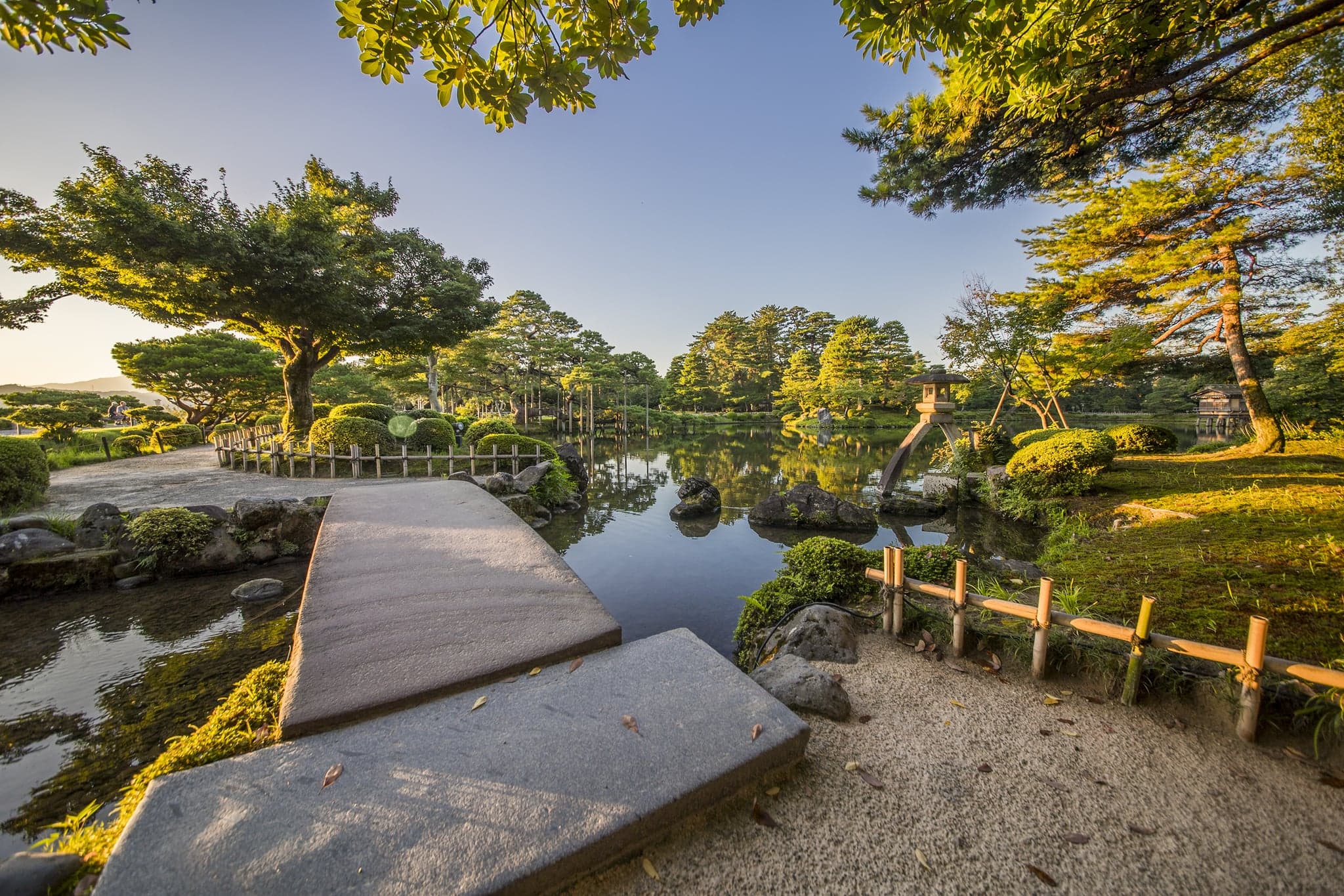 A pond with a stone path, pine trees and a low stone bridge in Kenrokuen garden in soft light