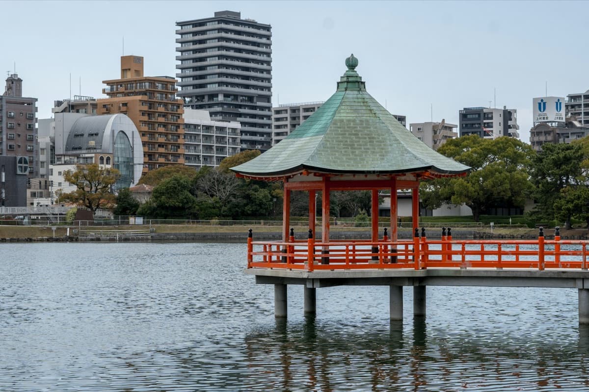 The red and green Ukimido pavilion standing on the water of Ohori Park's pond with city buildings behind
