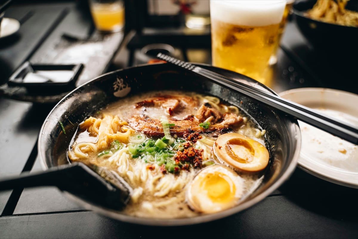 A bowl of tonkotsu ramen with slices of chashu pork, a soft-boiled egg and green onions, with a glass of beer behind