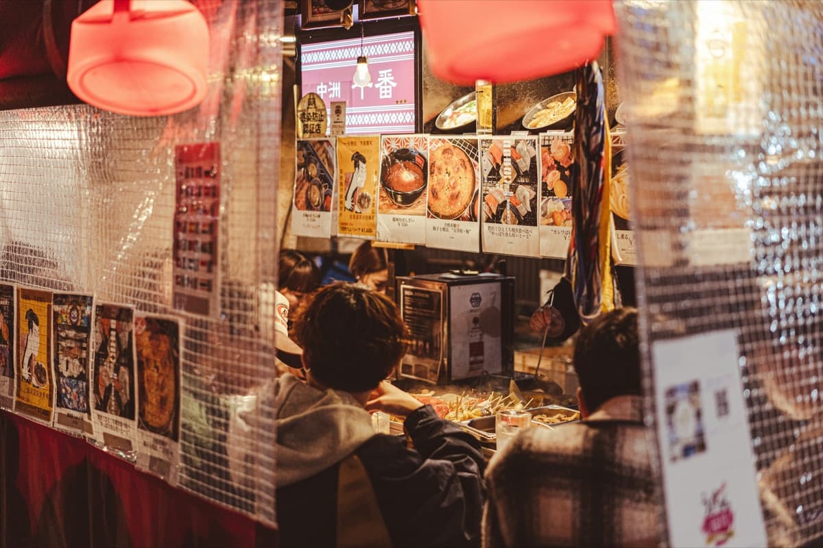 Customers sitting at the counter inside a yatai stall with menus and photos pinned to the plastic walls