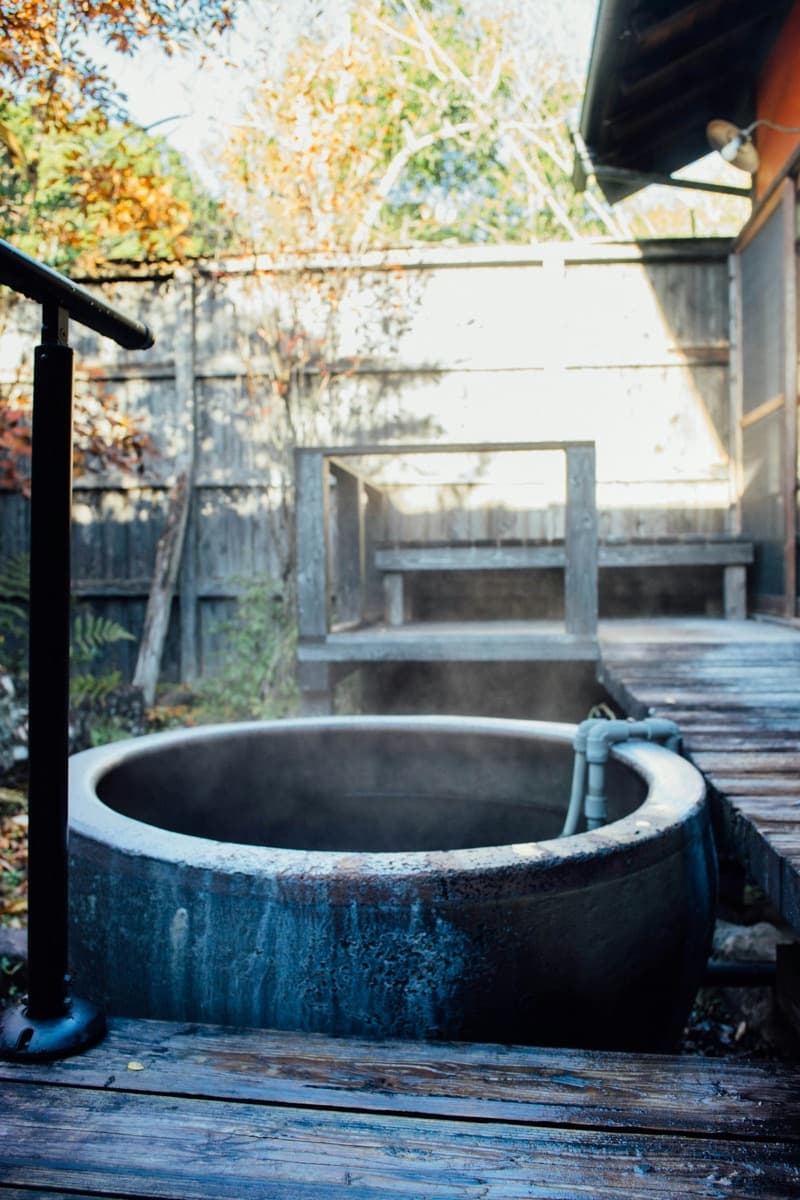 A steaming stone tub filled with hot spring water on a wooden deck, with a bamboo fence and autumn leaves behind it