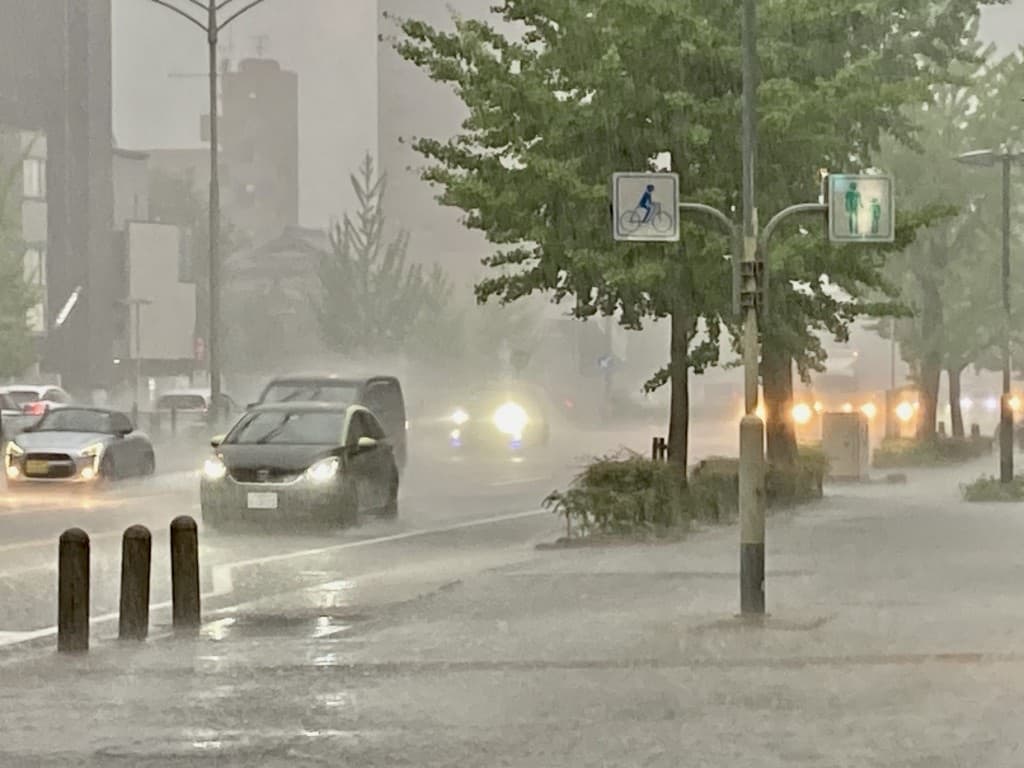 A Japanese city street under torrential rain, with cars driving through standing water during a sudden downpour