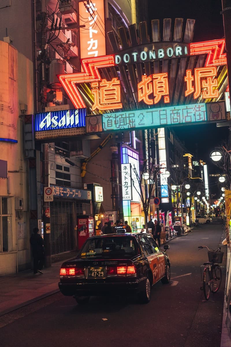 El letrero de entrada de Dotonbori iluminado en rojo y neón de noche, con un taxi en la calle