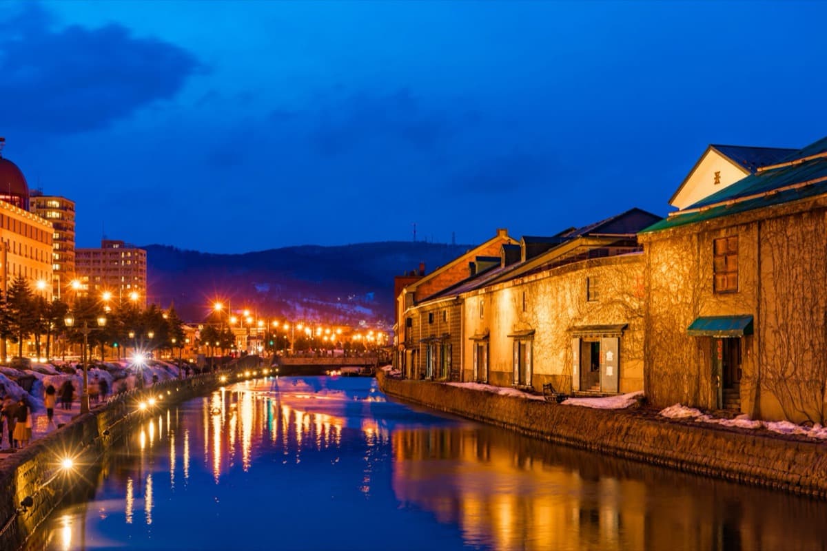 The Otaru canal in Hokkaido at night in winter, with warm street lamps reflected in the water and snow along the stone warehouse embankment
