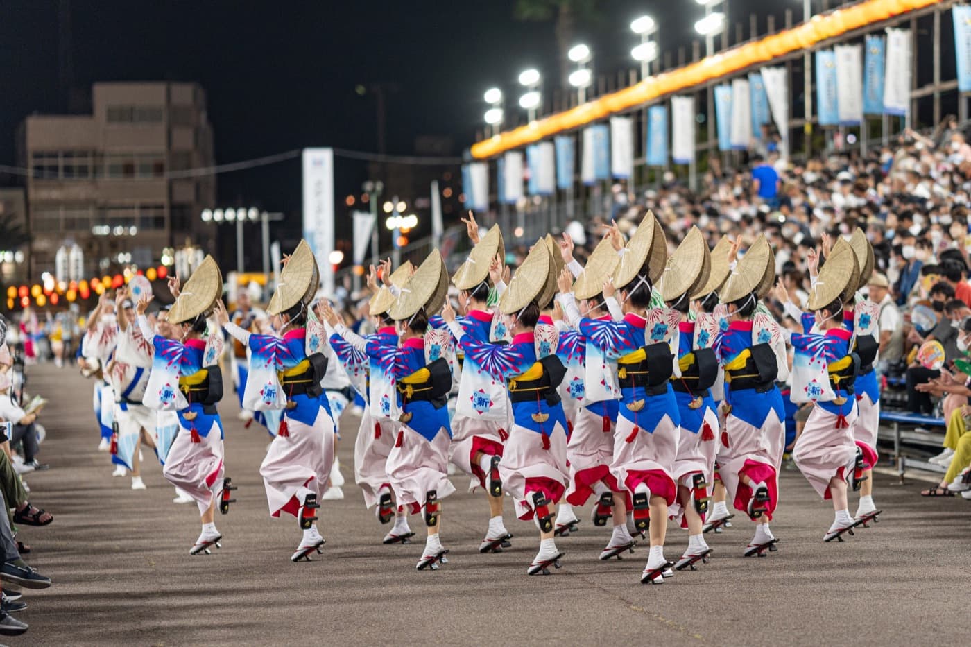 Awa-Odori-Tänzer mit Strohhüten in blau-weißen Yukata treten nachts vor einer voll besetzten Tribüne auf
