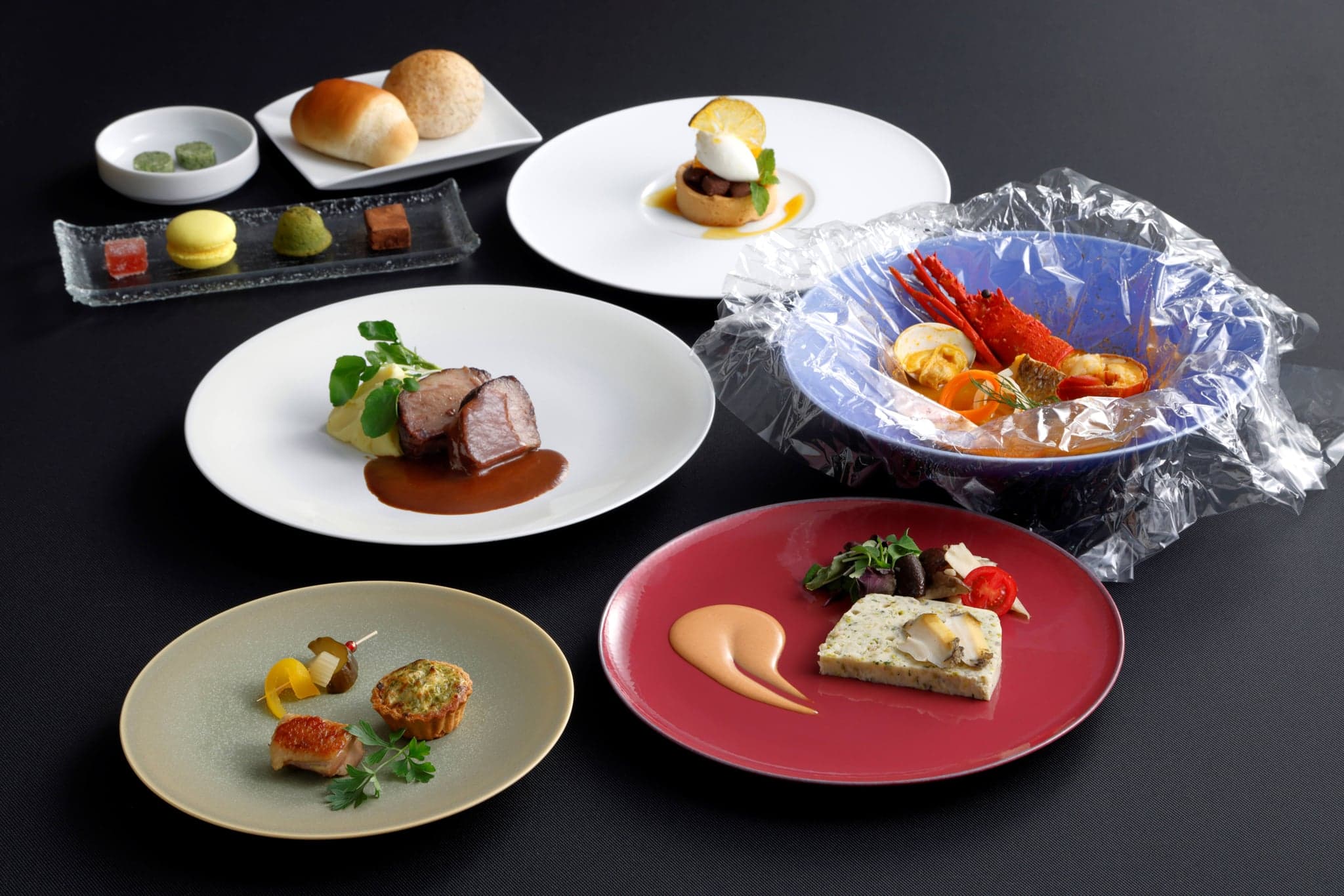 The French Course of Les Saveurs Shima laid out on a dark table: Ise lobster and clams in a blue bowl, abalone terrine, roast beef with sauce, bread, petits fours and a tart