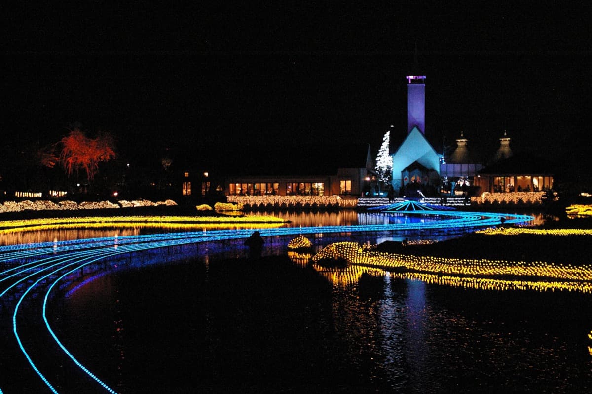 The Nabana no Sato chapel lit in white and blue at night, with curving lines of light on the pond in front