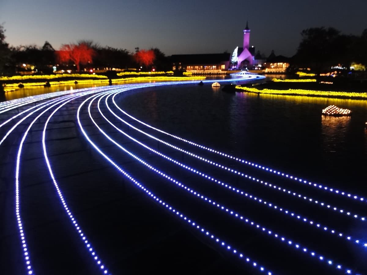 Curving lines of white light across the pond at dusk, with lit trees and the chapel tower in the distance