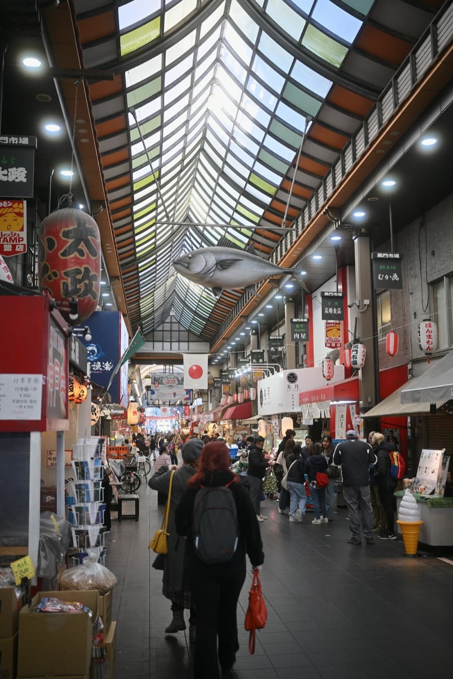 Gente caminando bajo el techo acristalado del mercado Kuromon junto a los puestos, con un pez de adorno colgado