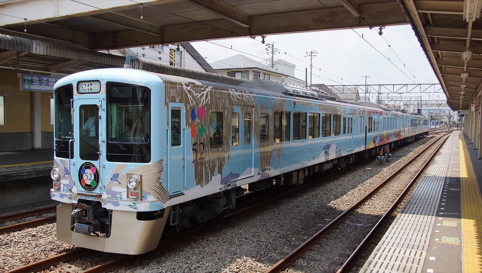 The pale blue 52 Seats of Happiness restaurant train of Seibu Railway, decorated with tree and cloud motifs, standing at a suburban station platform