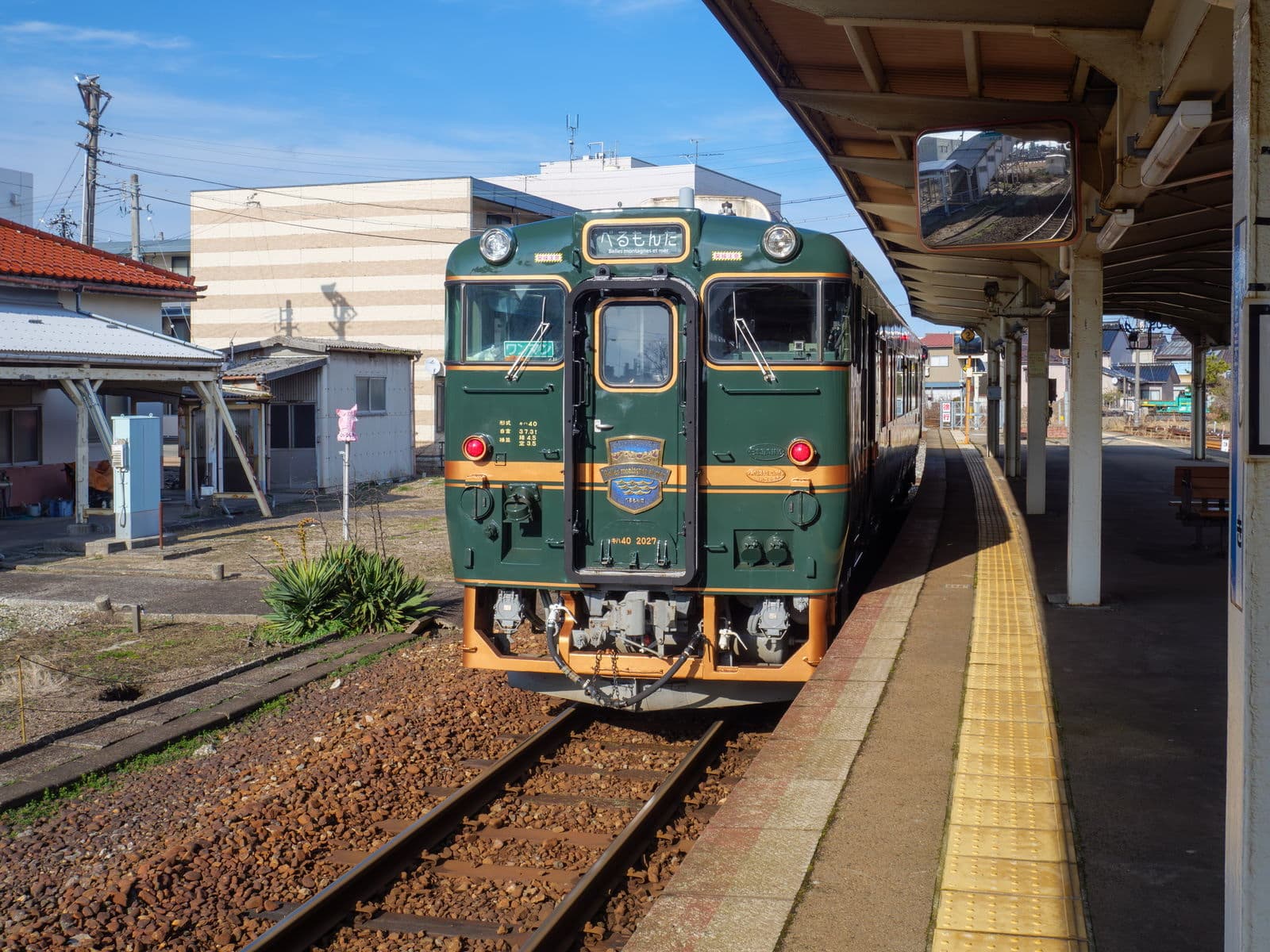 The dark-green Berumonta sightseeing railcar standing at a small station platform in Toyama under a blue sky