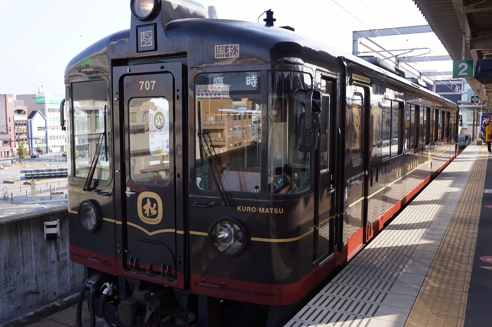 The black and red Tango Kuromatsu railcar of the Kyoto Tango Railway at a station platform, with the Kuro-Matsu pine emblem on its nose