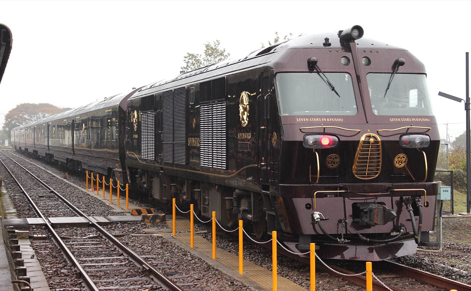 The dark maroon Seven Stars in Kyushu cruise train with gold emblems standing at Aso Station on a wet day