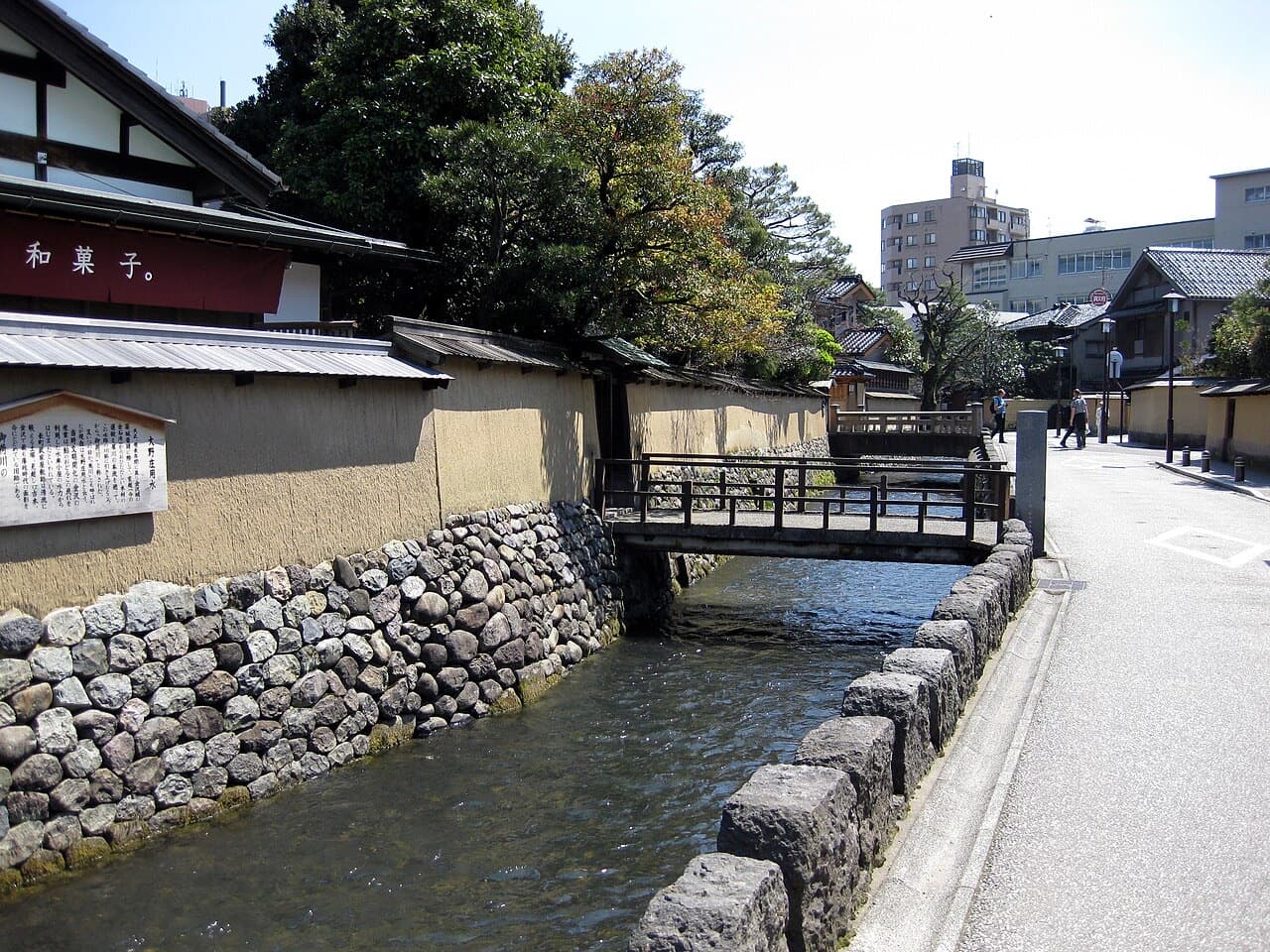 A narrow canal running beside a stone wall and a small wooden bridge in the Nagamachi district
