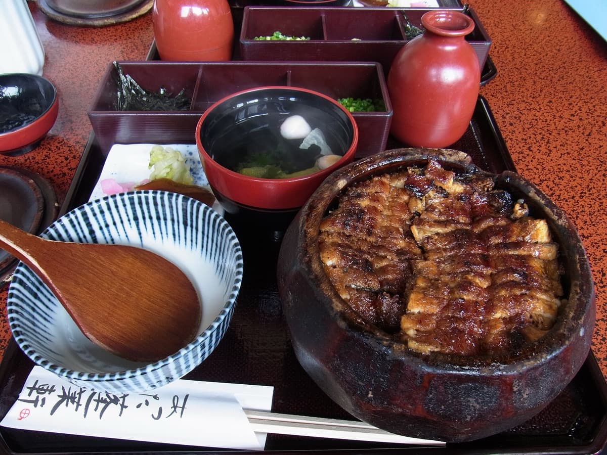 A hitsumabushi set with grilled eel over rice in a round wooden tub, condiments, soup and a pot of broth