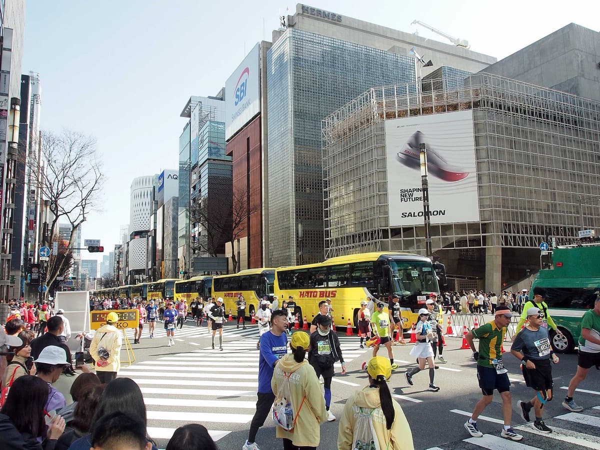 Runners passing the Sukiyabashi crossing in Ginza, with a long line of yellow Hato buses parked along the course