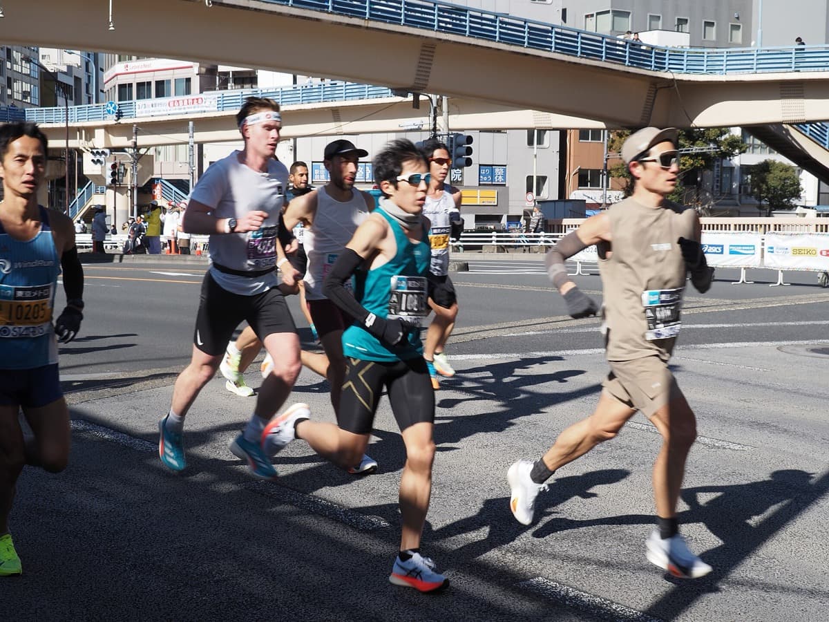 A group of marathon runners in the early kilometers passing under a pedestrian bridge on a sunny day