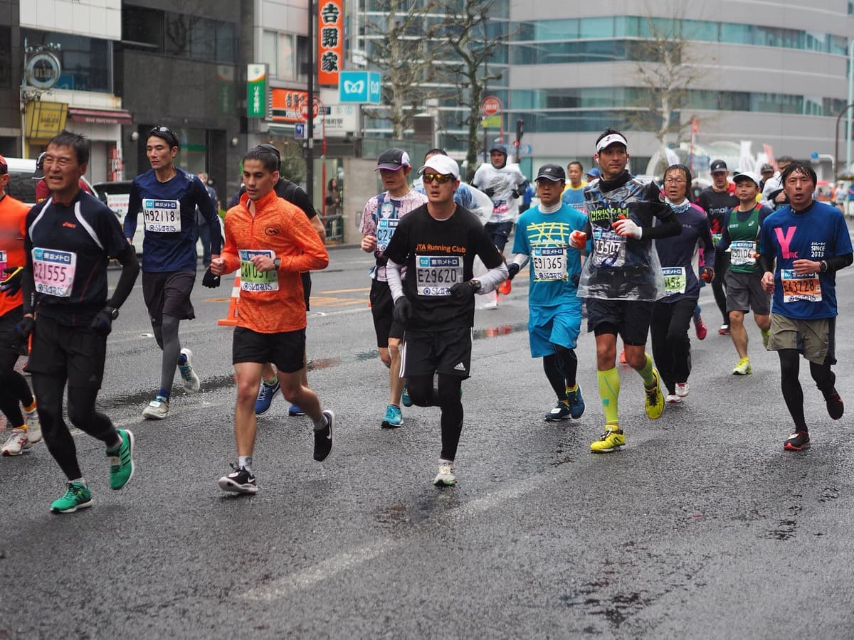 Marathon runners in gloves, jackets and ponchos running on a wet Tokyo street