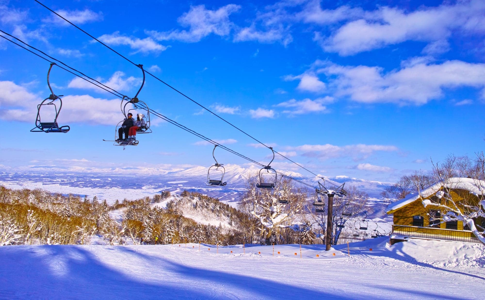 A chairlift carrying two skiers over a groomed Hokkaido slope with a snow-covered plain and mountains beyond