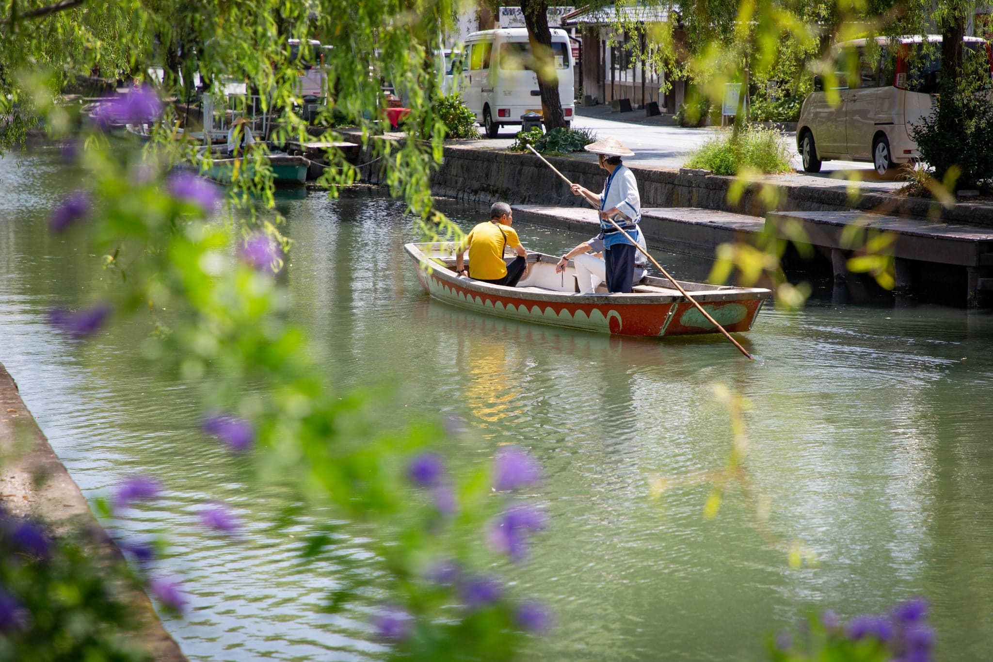 A boatman in a straw hat poling a flat punt with passengers along a willow-lined canal in Yanagawa