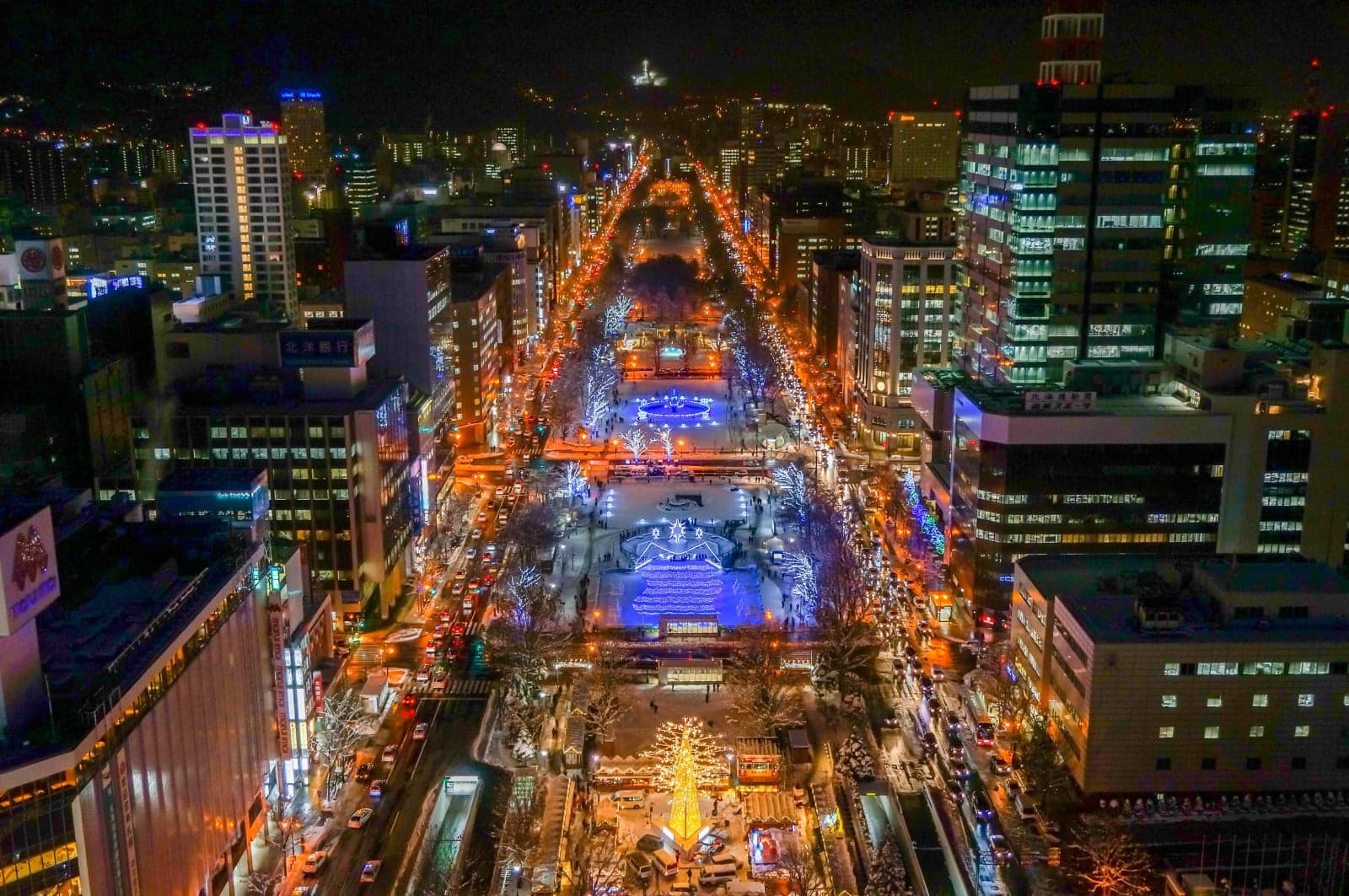 Odori Park lit up at night during the Sapporo Snow Festival, seen from above, with snow sculptures glowing along the park