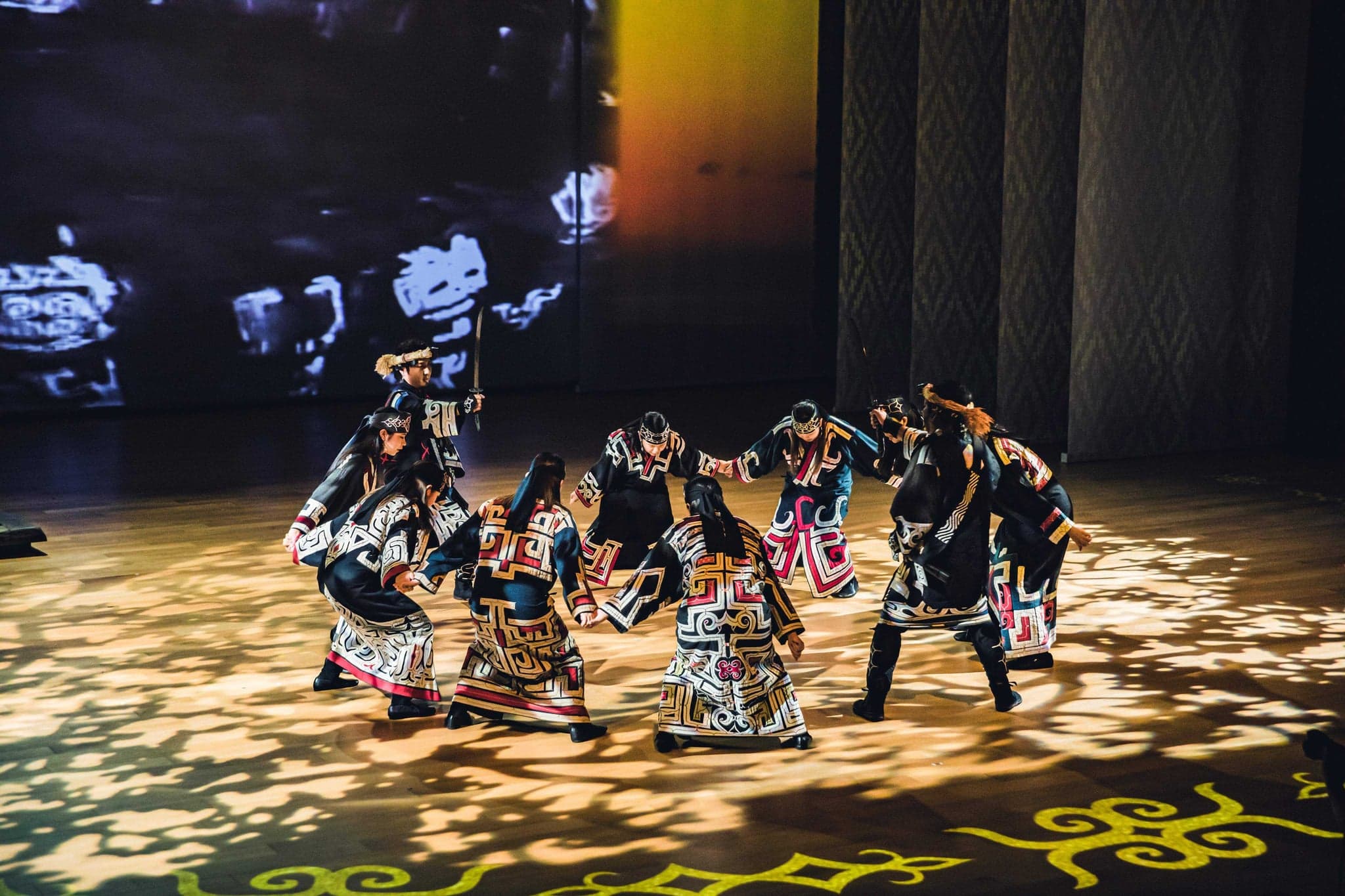 Ainu performers in patterned robes dancing in a circle on a dark stage at Upopoy