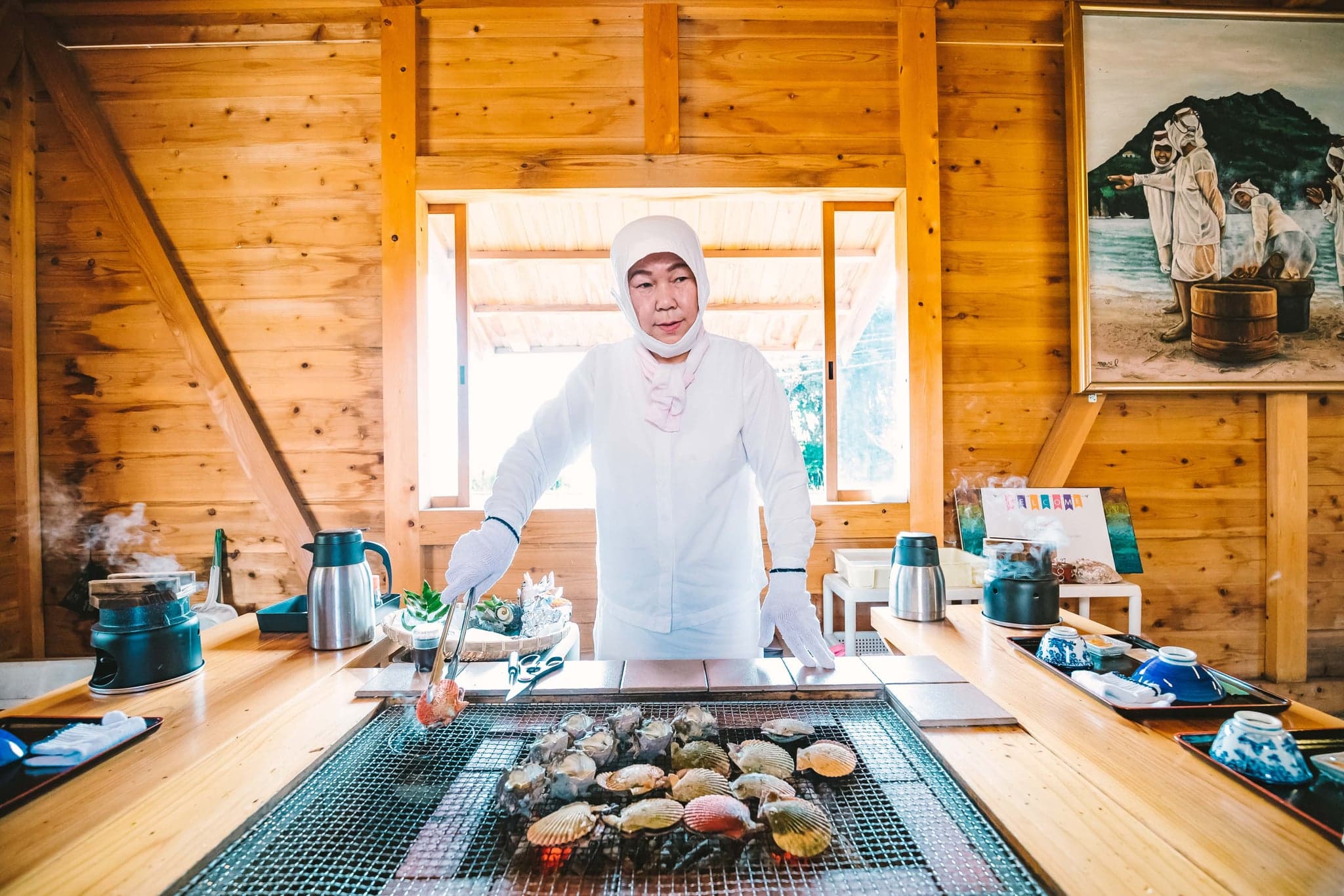 An ama diver in a white hood grilling scallops and shellfish over charcoal inside a wooden ama hut on the Shima coast