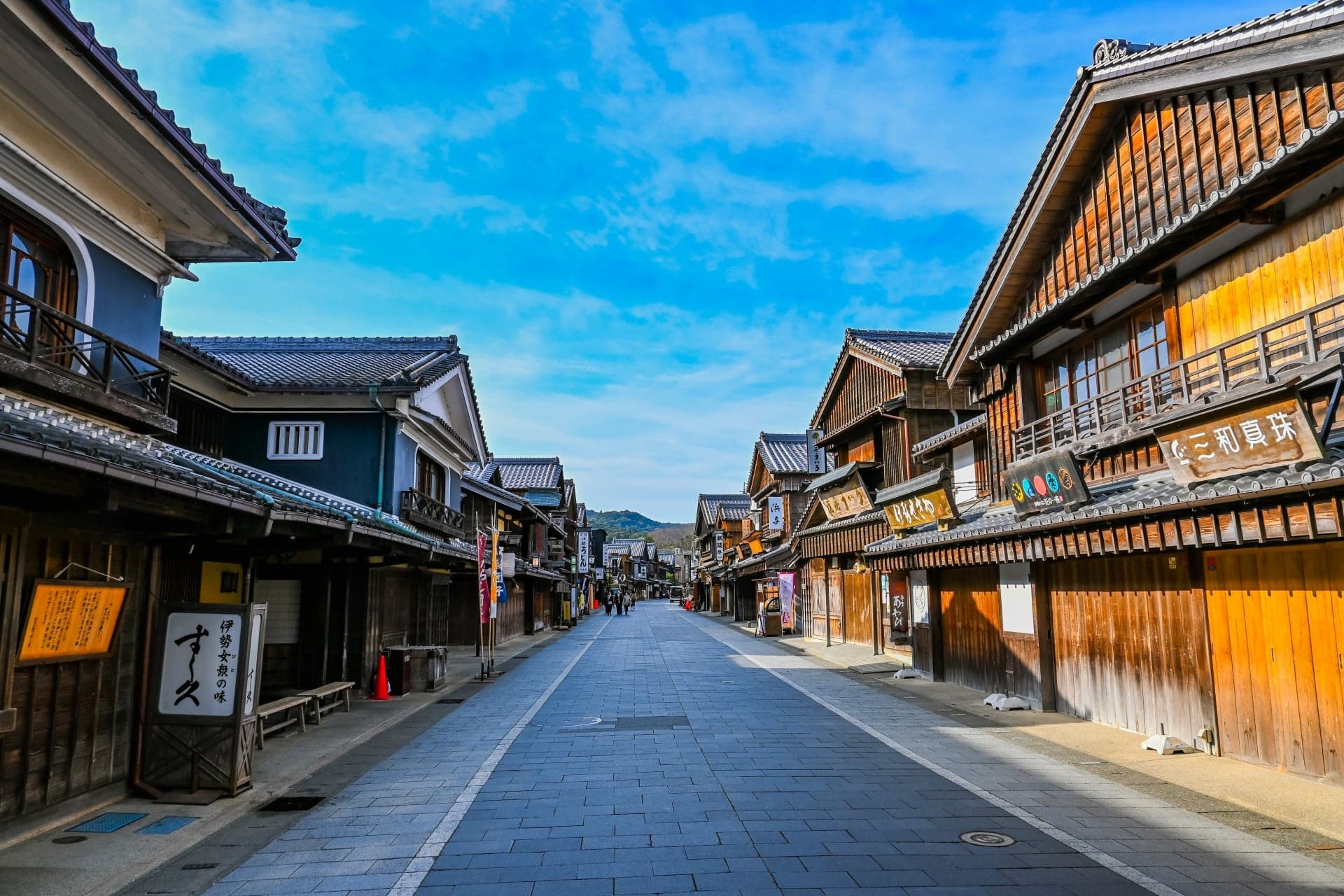 Traditional wooden merchant houses with hanging shop signs along the stone-paved street of Okage Yokocho in Ise under a blue sky