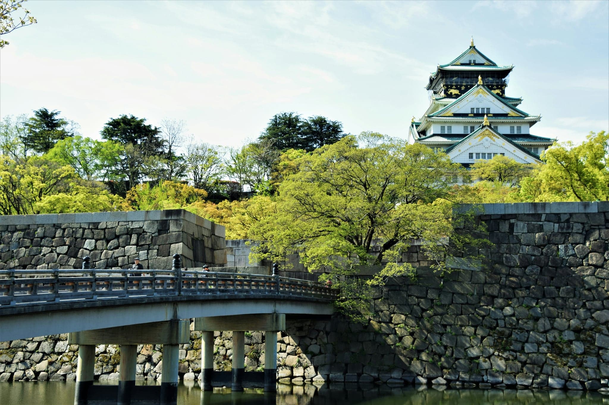 Osaka Castle's main tower above green trees, stone walls and a moat bridge
