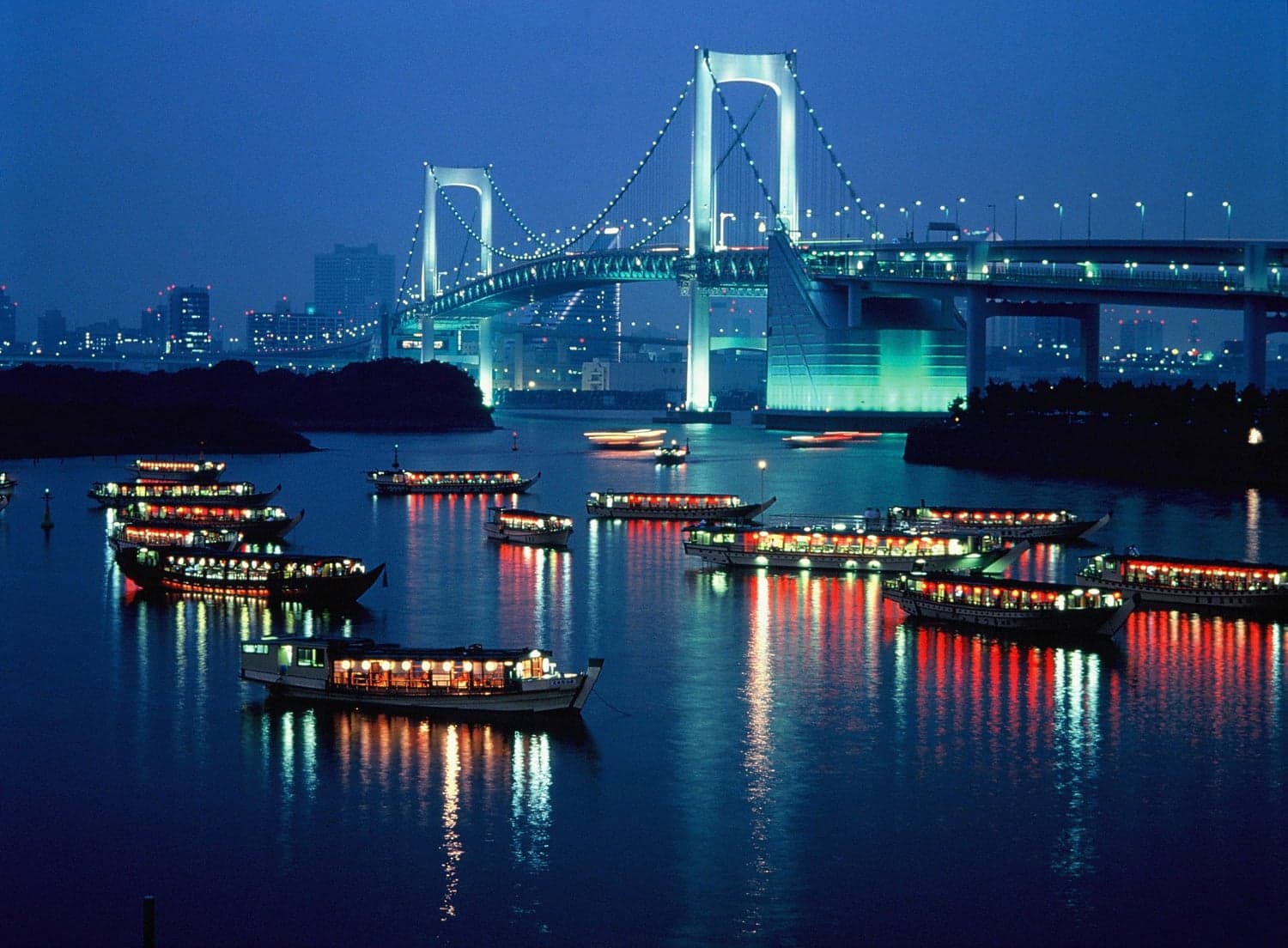 Rainbow Bridge lit up at night over Tokyo Bay with lantern-lit yakatabune boats on the water
